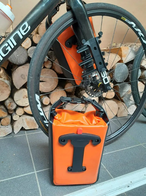 Sacoche de fourche vélo orange posée au sol, dos et fixation en T visibles, support monté sur la fourche