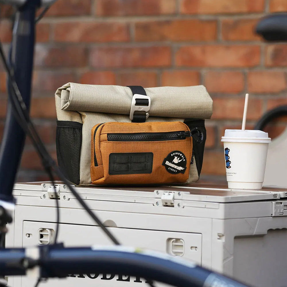 Sacoche de guidon beige avec poche frontale caramel posee sur une surface pres d un velo et d un cafe, devant un mur de briques.