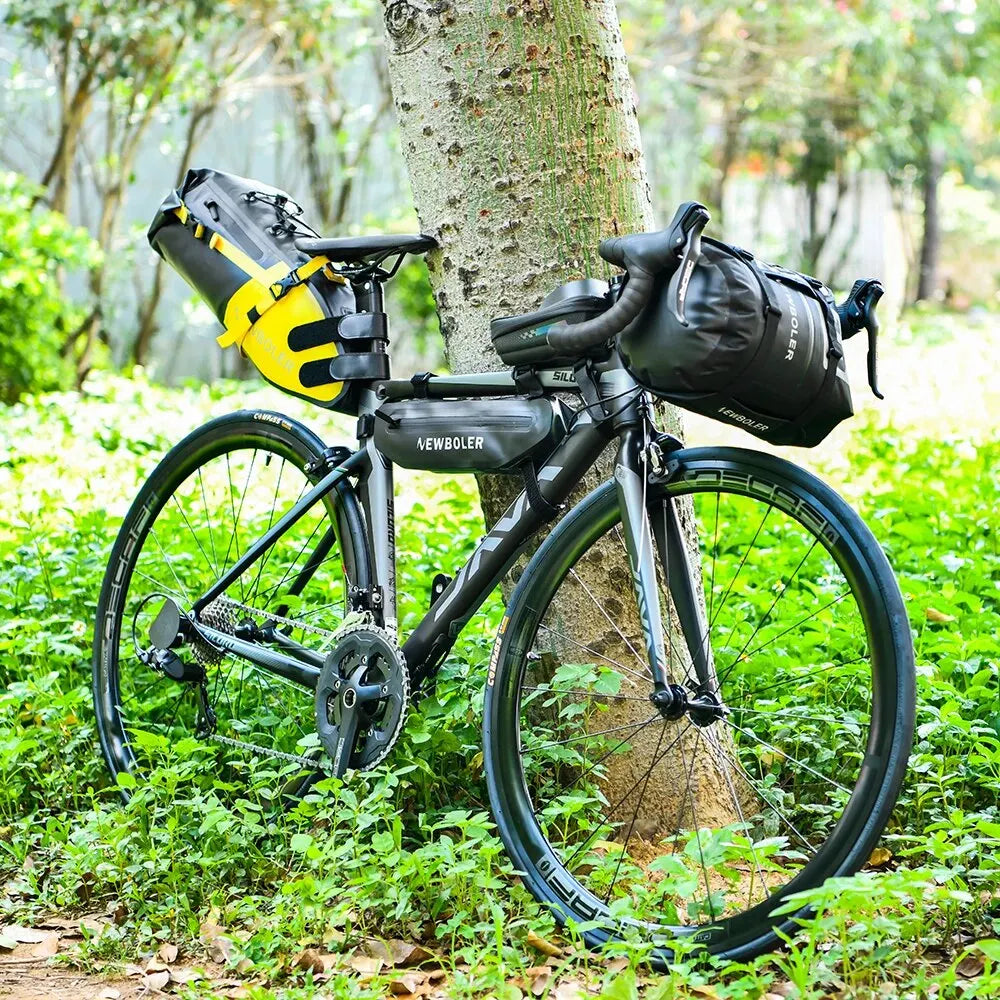 Velo de route noir equipe d'une sacoche de selle jaune et noire, d'une sacoche de guidon et d'une sacoche de cadre, appuye contre un arbre en foret.
