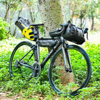 Velo de route noir equipe d'une sacoche de selle jaune et noire, d'une sacoche de guidon et d'une sacoche de cadre, appuye contre un arbre en foret.